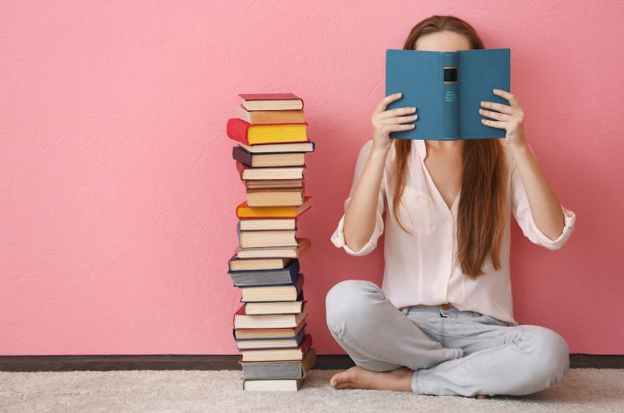 Woman sitting on a floor and holding book in front of face on pink background